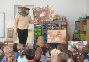 Pszczelarz prezentuje dzieciom obraz z rojem pszczół. W tle różne widać obraz ze słoikami z miodem.
